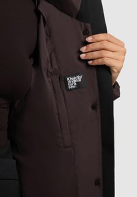 Bruine gewatteerde jas met zwarte accenten, voorzien van een gestructureerde binnenvoering, een logopatch en drukknoopsluitingen langs de opening.