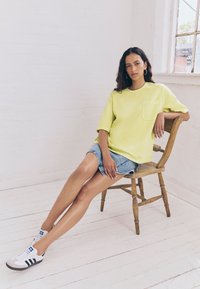 Maglietta gialla oversize con taschino frontale, abbinata a pantaloncini di jeans. Il modello è seduto su una sedia di legno, indossando sneakers bianche con strisce nere.