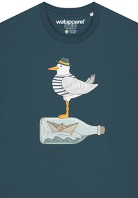 T-shirt turquoise avec un goéland portant une chemise rayée et un chapeau, se tenant sur une bouteille en verre avec un bateau en papier à l'intérieur et des vagues en dessous.