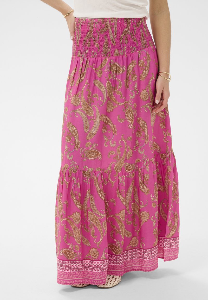 Žena oblečená v dlhej, splývavej fuchsiovej sukni s hnedými paisley a kvetinovými vzormi a viacvrstvovým lemom, v kombinácii s bielym topom.