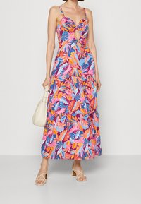 Robe maxi avec un motif floral vibrant en rose, bleu et jaune, dotée d'un détail noué à l'avant et de fines bretelles. Texture de tissu lisse.