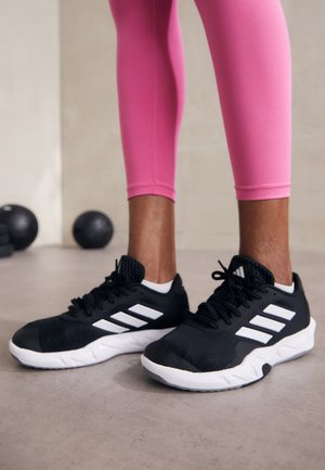 Persona con mallas rosa brillante y zapatillas negras Adidas con rayas blancas, de pie en interiores con balones medicinales negros al fondo.