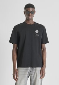 Camiseta de algodón negra de manga corta, con cuello redondo, que presenta un gráfico de un tigre blanco y texto en el lado izquierdo del pecho. Llevada con gafas de sol oscuras.