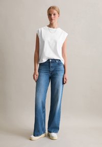 Weißes ärmelloses Baumwolltop kombiniert mit hellblauen weit geschnittenen Jeans. Das Modell trägt weiße Sneakers. Einfaches Design ohne sichtbare Muster oder Akzente.