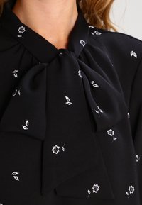 Blouse noire avec un col à nouer, motifs floraux blancs délicats et texture de tissu lisse, conçue pour une silhouette ajustée.