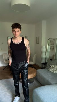 Camisola preta sem mangas e calças pretas brilhantes de couro ecológico, com um corte ajustado, usadas com ténis brancos. Fundo simples com decoração mínima.