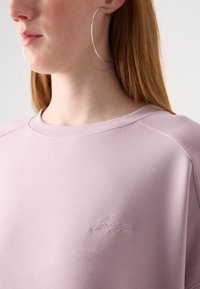 Světle růžový sweatshirt s texturovanou tkaninou, širokým výstřihem a vyšitým logem na hrudi. Má uvolněný, neformální design.