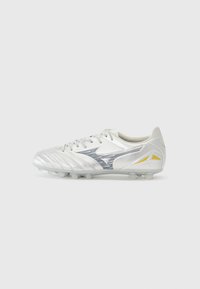 Crampons de football blancs avec des accents argentés, présentant une tige texturée, un système de laçage et des crampons inférieurs translucides. Logo et lignes de design remarquables.