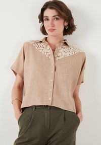 Bež lühikesleeve blouse, millel on heegeldatud pitsist yoke, nööbiga esiosa disain ja mugav lõige. Valmistatud tekstuurist kangast, millel on peen sära.