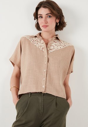Beige short-sleeve blouse with a crochet lace yoke, button-front design, and a relaxed fit. Made of textured fabric with a subtle sheen.