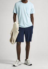 Lyseblå t-shirt med lomme, navy cargo shorts, beige hættetrøje over armen, og grå sneakers med hvide detaljer. Afslappet outfit.