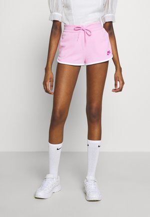 Person iført lyserøde Nike-shorts med hvid kant, hvide Nike-strømper, hvide sneakers og en hvid kortærmet top, står mod en ensfarvet baggrund.