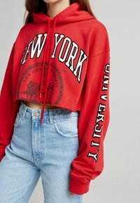 Sudadera roja corta con capucha ajustable, que presenta "NUEVA YORK" en letras blancas en el pecho y "UNIVERSIDAD" en la manga, combinada con jeans de color azul claro.