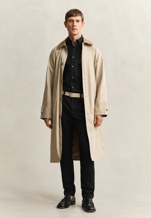 Man draagt een beige trenchcoat over een zwart shirt en zwarte broek, met een beige riem en zwarte loafers, staand tegen een effen achtergrond.