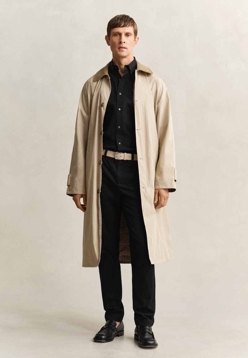 Homme portant un trench-coat beige par-dessus une chemise noire et un pantalon noir, avec une ceinture beige et des mocassins noirs, debout devant un fond uni.