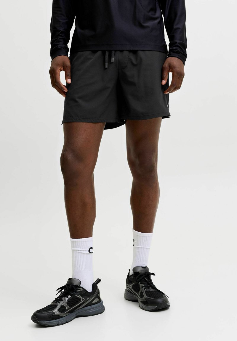 Homme portant un short de sport noir, un t-shirt noir à manches longues, des chaussettes blanches montantes et des chaussures de course noires, debout sur un fond blanc.
