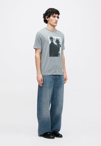 Γκρι βαμβακερό T-shirt με ασπρόμαυρη γραφική εκτύπωση στο μπροστινό μέρος. Συνδυασμένο με μπλε τζιν παντελόνι φαρδιάς γραμμής και μαύρα παπούτσια.