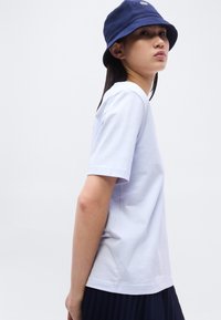 Jeune personne portant une chemise blanche à manches courtes, un chapeau seau bleu marine et une jupe plissée foncée, vue de profil sur un fond clair uni.