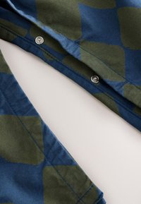 Tissu en coton avec une base bleue ornée de grandes formes abstraites vertes. Détail de fermeture à pression et surpiqûres contrastantes visibles.