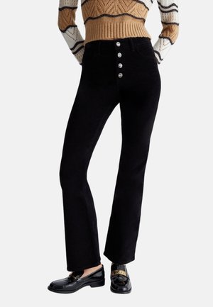 Woman wearing black high-waist pants with four visible buttons and black patent leather loafers with gold detail.