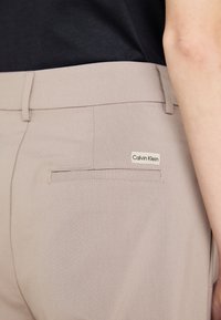 Beige kohandatud püksid, millel on sile tekstuur, tagaküljel klapiga tasku ja väike Calvin Kleini silt.