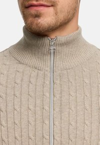 Beige kabelstrikket genser med en høy ribbestrikket krage, som har en glidelås foran. Teksturen er myk, og mønsteret består av sammenvevde kabler.