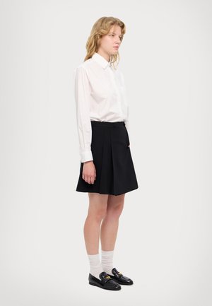 Camicia bianca con bottoni e maniche lunghe, gonna plissettata nera, calze bianche e mocassini neri con un accento dorato. Design semplice, colori classici.