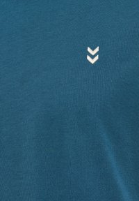 Blå bomullströja med en strukturerad tyg och en liten vit chevronlogotyp på framsidan. Designen är enkel och minimalistisk.