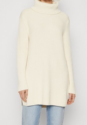 Femme portant un pull à col roulé en maille côtelée crème avec manches longues et fentes sur les côtés, associé à un jean bleu clair.