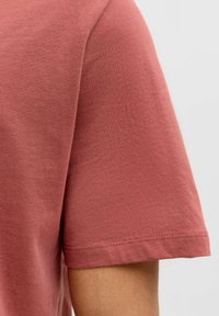 Primer plano del brazo de una persona que lleva una camiseta de algodón de color rosa empolvado, de manga corta y sin estampado, contra un fondo blanco.