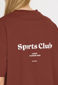 Bordeauxfärgad bomulls-T-shirt med korta ärmar, med vit text: "Sports Club" och "Luxury Athleisure Wear" på baksidan, minimalistisk design.