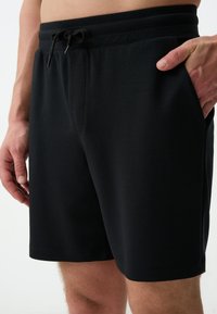 Shorts noirs en tissu lisse, dotés d'une taille élastique avec cordon de serrage et de poches latérales. Simples, sans motifs ni ornements.