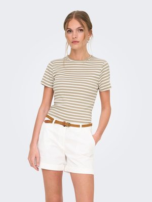 Vrouw draagt beige-wit gestreept kort mouwen topje, witte shorts met opgerolde zomen en een gevlochten bruine riem, staand met één hand in de zak.