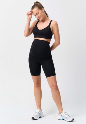 Woman standing and smiling, wearing black sports bra and high-waisted black biker shorts with white athletic shoes against a plain background.