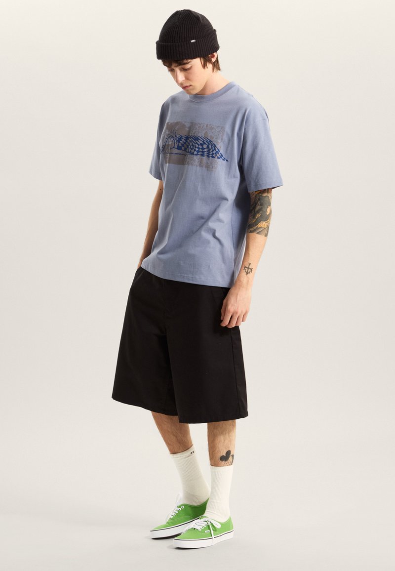 Lichtblauw katoenen T-shirt met grafisch ontwerp, zwarte korte broek, witte sportsokken en groene sneakers. Model draagt een zwarte gebreide muts.