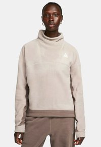 Felpa in pile beige chiaro con colletto alto, polsini a coste e un logo triangolare sul petto, abbinata a pantaloni coordinati.