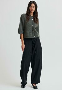 Tmavo zelený krátky pletený kardigán s krátkymi rukávmi a veľkými gombíkmi, skombinovaný s čiernymi nohavicami s vysokým pásom a širokými nohavicami a špicatými topánkami.