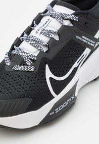 Chaussure de trail running noire avec une tige en mesh respirant, accents blancs, semelle texturée et marquage distinctif Nike ZoomX. Présente des lacets à motifs.