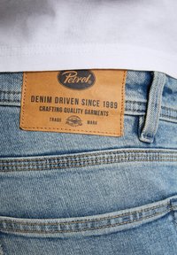 Vaqueros de mezclilla con un lavado claro que presentan una etiqueta de cuero marrón con la inscripción "Petrol, Denim Driven Since 1989, Crafting Quality Garments."