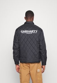 Černá prošívaná bunda s límcem a bílým nápisem "CARHARTT" na zádech. Hladká látka s diamantovým prošíváním a manžetami na knoflíky.