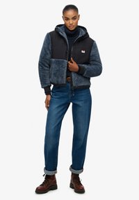 Czarna i granatowa kurtka z podszewką z polaru, z kieszeniami na zamek, kapturem i ściągaczami; w połączeniu z podwiniętymi dżinsami i brązowymi butami.