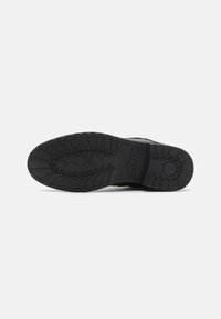Semelle de chaussure noire avec un motif de crampons profond, des points d'adhérence circulaires et la marque "bugatti" embossée près du talon.