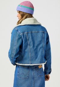 Jeansowa kurtka z jasnym futrzanym kołnierzem, o kroju w stylu cropped, w klasycznym niebieskim kolorze oraz widocznymi detalami szycia.