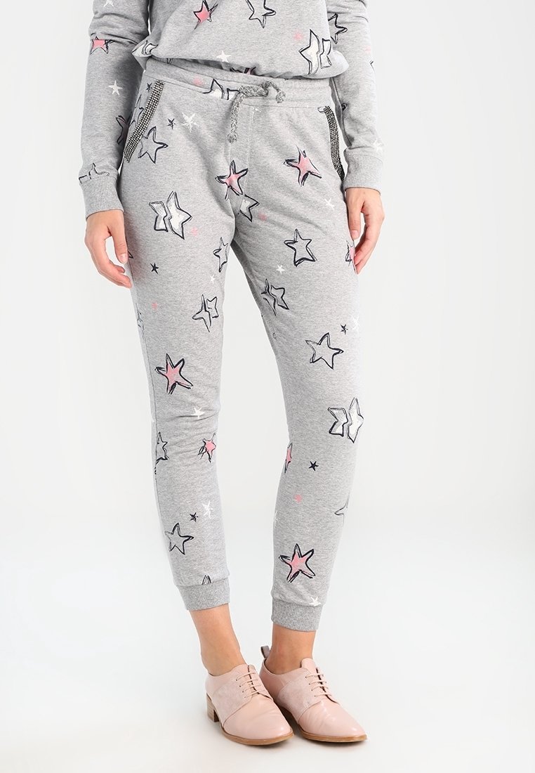 Person trägt graue Jogginghosen mit pinken und schwarzen Sternmustern sowie einen passenden Pullover, kombiniert mit hellrosa Oxford-Schuhen.