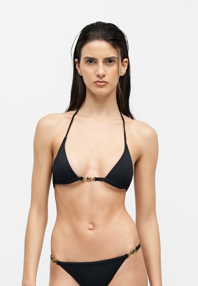 Jeune femme aux cheveux mouillés portant un bikini deux pièces noir avec des accents d'anneaux dorés, face à un fond clair uni.