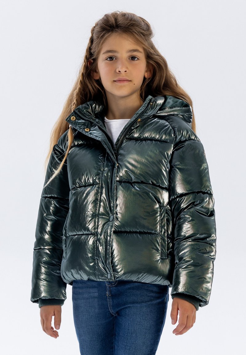 Grön metallisk puffer-jacka med huva, som har en glänsande, vadderad yta, tryckknappar och ribbade ärmslut. Bärs lager på lager över en vit skjorta.