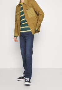 Kvadret brun jacka med två bröstfickor, en grön och gulrandig långärmad skjorta under, mörkblå jeans och svarta sneakers.