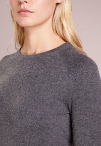 Grå stickad tröja med rundad halsringning och raglanärmar, med en mjuk textur och en slät yta.