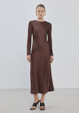 Junge Frau steht in einem braunen, enganliegenden Midi-Kleid mit langen Ärmeln, schwarzen High-Heeled Sandalen und goldenen geometrischen Ohrringen.