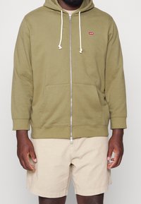 Hombre vistiendo una sudadera con cremallera de color verde oliva con cordones blancos y pantalones cortos beige, sobre un fondo liso.
